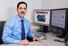Dr. Khalid Shah at his desk in the CSTI laboratory, with research images displayed on two monitors behind him.